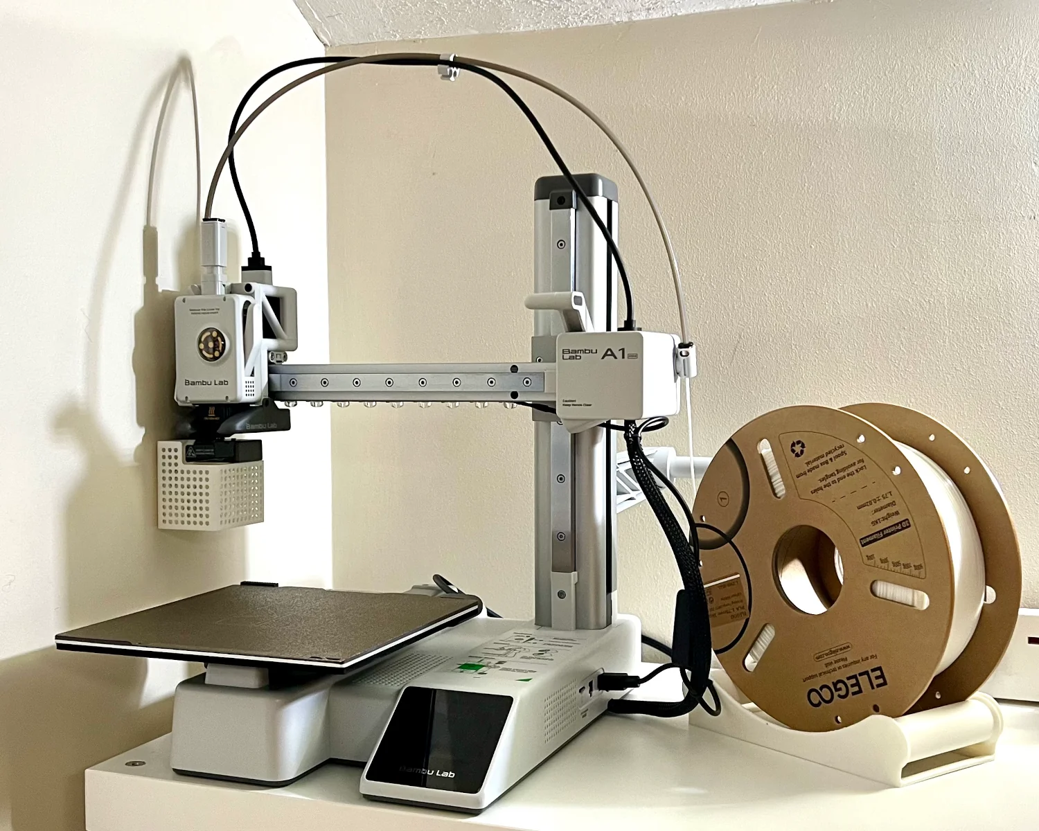 A Bambu Lab A1 Mini next to a roll of White Elegoo PLA filament sitting on a pair of rollers.